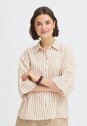Mladá žena s krátkymi tmavými vlasmi nosí zlaté kruhové náušnice, béžovú a bielo pruhovanú košeľu na gombíky, s prekríženými rukami a miernym úsmevom.