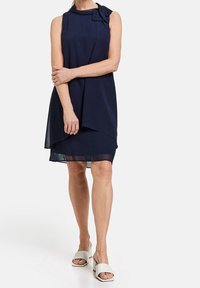 Vestido sin mangas de color azul marino con detalle de lazo en el hombro, diseño en capas y textura suave. Lleva sandalias de deslizar blancas.