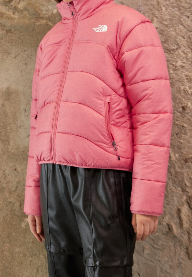 Giacca a vento rosa con colletto alto, chiusura con zip e tasche laterali. Presenta motivi di cucitura e un logo sul petto. Abbinata a pantaloni neri.
