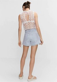 Bílý háčkovaný top s otevřeným zadním dílem a detailem na zavazování, doplněný modro-bílými proužkovanými šortkami. Modelka nosí béžové slip-on sandály.