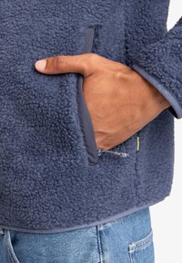 Navy fleece pullover with a textured, fluffy exterior, featuring a front pocket, zipper accent, and elastic cuffs in a contrasting material.