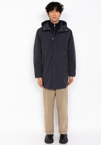 Parka impermeable azul marino con cremallera frontal, dos bolsillos laterales con cremallera, capucha y un corte relajado, llevada sobre pantalones beige y zapatos negros.