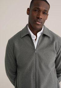 Mężczyzna noszący szarą kurtkę zapinaną na zamek, na białą koszulę z kołnierzykiem, stojący na tle jednolitego jasnego tła, z neutralnym wyrazem twarzy.