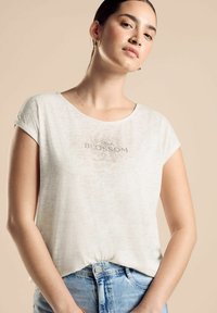 Bílý zkrácený tričko s kulatým výstřihem a krátkými rukávy. Obsahuje strukturovaný vzor a nápis "BLOSSOM" vytištěný na přední straně.