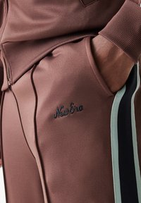 Μαρόν φόρμα με ριμπ λεπτομέρειες και κεντημένο λογότυπο "New Era". Οι λωρίδες στο πλάι σε ανοιχτό πράσινο και μαύρο προσθέτουν λεπτομέρεια στο σχέδιο.