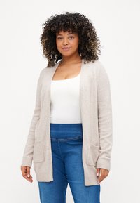 Light beige cardigan made of soft material, featuring an open front, ribbed cuffs, and two front pockets. Worn over a white top and blue jeans.