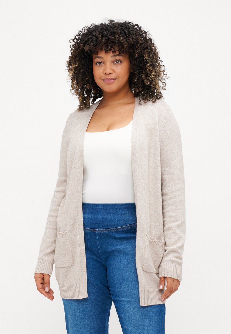 Light beige cardigan made of soft material, featuring an open front, ribbed cuffs, and two front pockets. Worn over a white top and blue jeans.