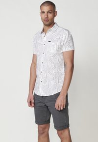 Camisa de manga corta con base blanca, patrón de hojas gris claro, botón en la parte delantera y pequeño logo en el pecho. Combinada con pantalones cortos grises remangados.