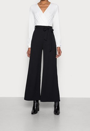 Haut blanc à manches longues enroulé avec un col en V, associé à un pantalon noir taille haute à jambes larges, ceinturé avec une ceinture à nouer. Bottines noires.