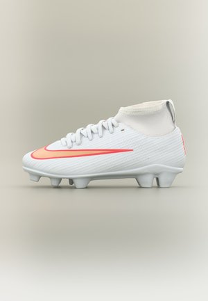 Bota de fútbol blanca con el logo rojo de Nike, cordones, cuello alto en el tobillo y tacos en la suela, mostrada sobre un fondo liso claro.