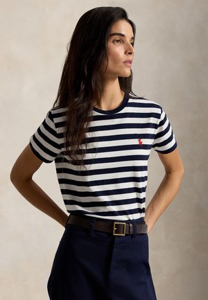 Femme aux longs cheveux foncés portant un t-shirt à manches courtes rayé bleu marine et blanc et un pantalon bleu marine avec une ceinture marron, regardant sur le côté.
