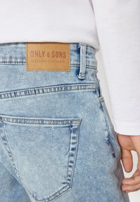 Ljust blå denimjeans med en läderlapp som visar "ONLY & SONS" vid midjan. Funktioner inkluderar sömmar och bakfickor.