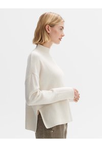 Heller Cremefarbener Pullover mit hohem Kragen, Rippenelementen, weiten Ärmeln und seitlichen Schlitzen, gefertigt aus einem weichen Material mit glatter Textur.