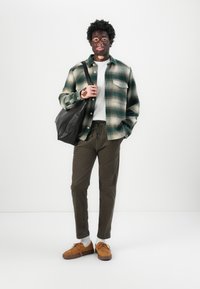 Geruite flanellen shirt in groen en room, witte t-shirt, olijfgroene broeken, tan suède schoenen en een zwarte leren tote bag.