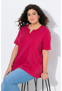 Top fucsia a maniche corte con scollatura a V, realizzato in tessuto morbido. Abbinato a jeans azzurri, modello seduto su uno sgabello nero.
