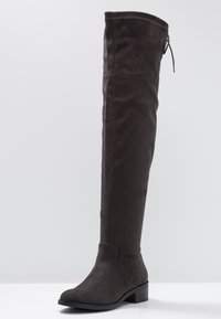 Botas sobre la rodilla en ante sintético de color marrón oscuro, con puntera redondeada y tacón bajo en bloque. Presentan una costura y un lazo ajustable en la parte superior.