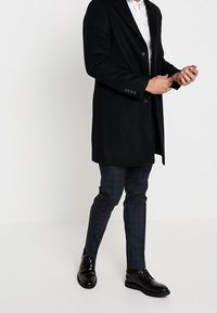 Zwarte jas met een getailleerde pasvorm, een eengrijpont ontwerp en twee knopen, gecombineerd met donker geruite broeken en gepolijste zwarte schoenen.