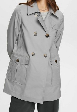 Femme portant un trench-coat gris clair à double boutonnage avec de grandes poches devant et des boutons foncés, associé à un pantalon foncé et un collier à chaîne.