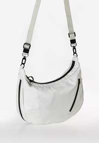 Weiße Nylon-Umhängetasche mit gewellter Form, ausgestattet mit schwarzen Reißverschlüssen, einem abnehmbaren Tragegurt und Metallakzenten. Glatte, weiche Textur.