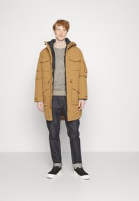 Doudoune beige matelassée avec plusieurs poches à l'avant, portée par-dessus un pull gris et un jean foncé, associée à des baskets noires.