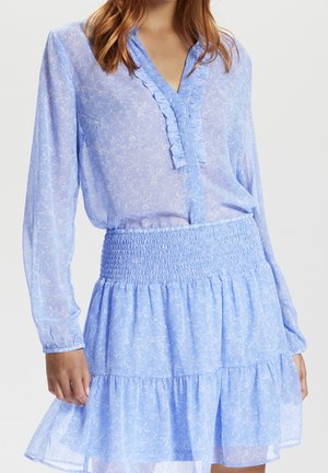 Vrouw draagt een lichtblauw doorschijnend bloesje met bloemenprint en ruches, gecombineerd met een bijpassende hoog getailleerde rok met lagen, tegen een effen achtergrond.