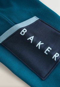 La tela azul verdoso presenta un parche de un oscuro navy con la palabra "BAKER" en letras blancas. El diseño combina dos texturas contrastantes.