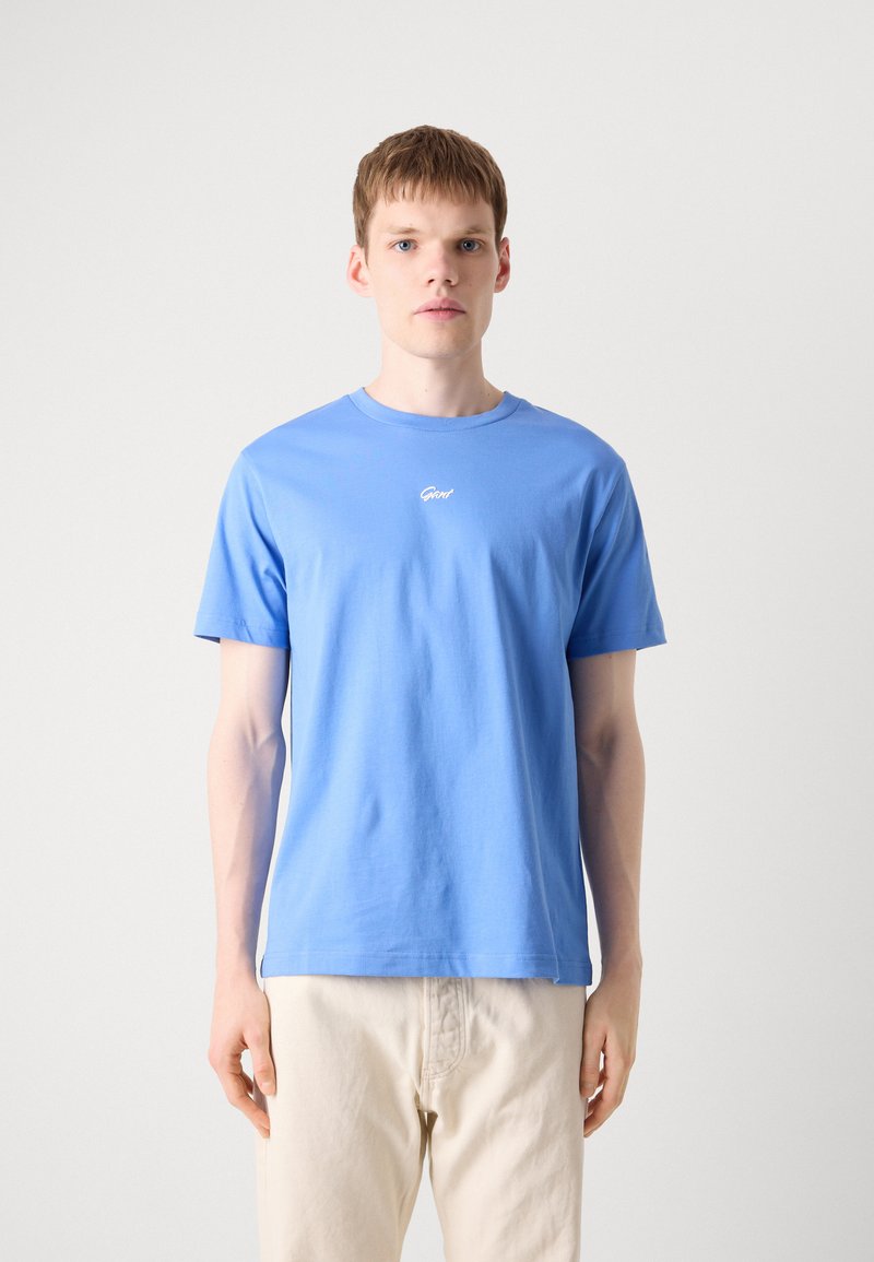 Niebieska bawełniana koszulka z okrągłym dekoltem, krótkimi rękawami i subtelnym białym logo na piersi, zestawiona z jasnymi spodniami.