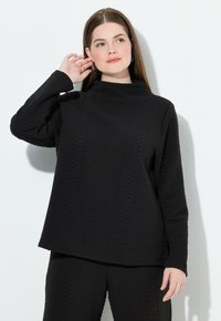 Młoda kobieta z długimi brązowymi włosami, ubrana w teksturowany czarny długi rękaw oraz pasujące spodnie, pozująca z ręką przy uchu na białym tle.