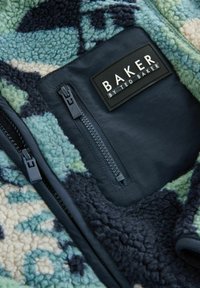 Vilnas jaka ar ziedu rakstu zaļā un tumši zilā toņos. Ir rāvējslēdzēja kabata ar melnas ādas etiķeti, uz kuras rakstīts "BAKER BY TED BAKER."