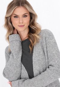 Giovane donna con capelli biondi mossi che indossa un cardigan grigio a maglia sopra un top scuro, in posa con una mano vicino al collo su uno sfondo bianco.