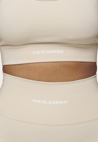 Beige sports bra and high-waisted leggings feature PEGAADOR logo. Smooth, stretchy material with a plain texture and fitted design.