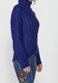 Pull à col roulé bleu tricoté, avec des motifs texturés et des poignets côtelés, associé à un jean brodé de motifs floraux sur la jambe.