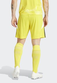 Heldere gele voetbaluniform met korte shorts, hoge sokken en witte voetbalschoenen. Kenmerken zijn de textuur van de stof en accenten aan de zijkant.