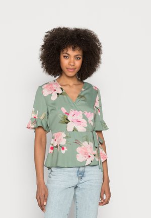Blusa verde com padrão floral, com mangas curtas e franzidas, apresentando padrões de flores rosa e brancas e um laço lateral. Tecido suave e leve.