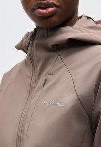 Gros plan sur une personne portant une veste taupe Columbia avec une fermeture éclair et une capuche, affichant le logo de l'entreprise sur la poitrine.