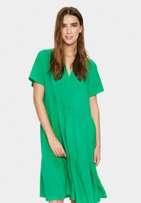 Rochie verde, până la genunchi, cu decolteu în v și mâneci scurte, având fusta din straturi. Fabricată dintr-un material ușor, cu un croi lejer și o textură netedă.