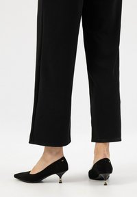 Černé semišové lodičky na vysokém podpatku s špičatou špičkou, zdobené zlatým akcentem na podpatku. Nosené s černými kalhotami, které ukazují elegantní design.