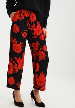 Vrouw die zwarte broek met wijde pijpen en felle rode bloemenprint draagt, zwarte trui en zwarte hoge hakken, staand met handen in de zakken.