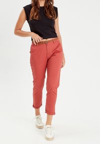 Haut noir sans manches, pantalon court rouge, ceinture tressée marron, et baskets blanches aux semelles beiges. Tissus simples et lisses avec revers roulés.