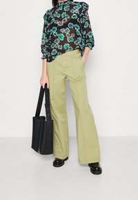 Blouse noire à motifs floraux avec manches à volants, associée à un pantalon large vert clair. Le mannequin porte un sac fourre-tout noir et des bottes noires épaisses.