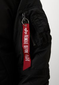 Manche de veste noire avec poche zippée et étiquette en tissu rouge attachée portant l'inscription "REMOVE BEFORE FLIGHT" en blanc.