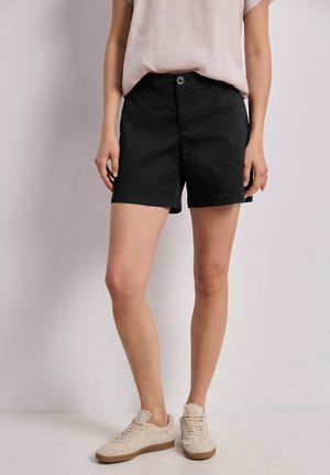 Person iført sorte knælange shorts, lysebeige kortærmet top og beige sneakers, der står op ad en almindelig væg.