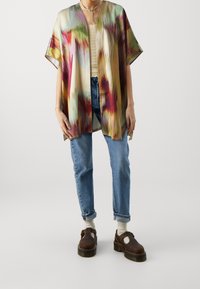 Lekki, wielokolorowy kimono o żywym, abstrakcyjnym wzorze, noszone na teksturowanej, off-white bluzce, zestawione z dżinsami i brązowymi butami.