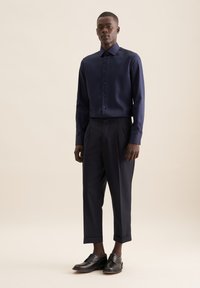 Camicia da uomo blu scuro con colletto a bottone, abbinata a pantaloni blu scuro plissettati e scarpe in pelle nere, che mostrano una vestibilità sartoriale e una texture liscia.