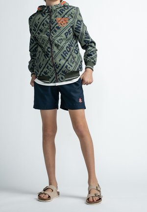 Zelená vzorovaná bunda s kapucí, zapínáním na zip a manžetovými rukávy, nošená přes bílé tričko, v kombinaci s námořnickými šortkami a béžovými sandály.