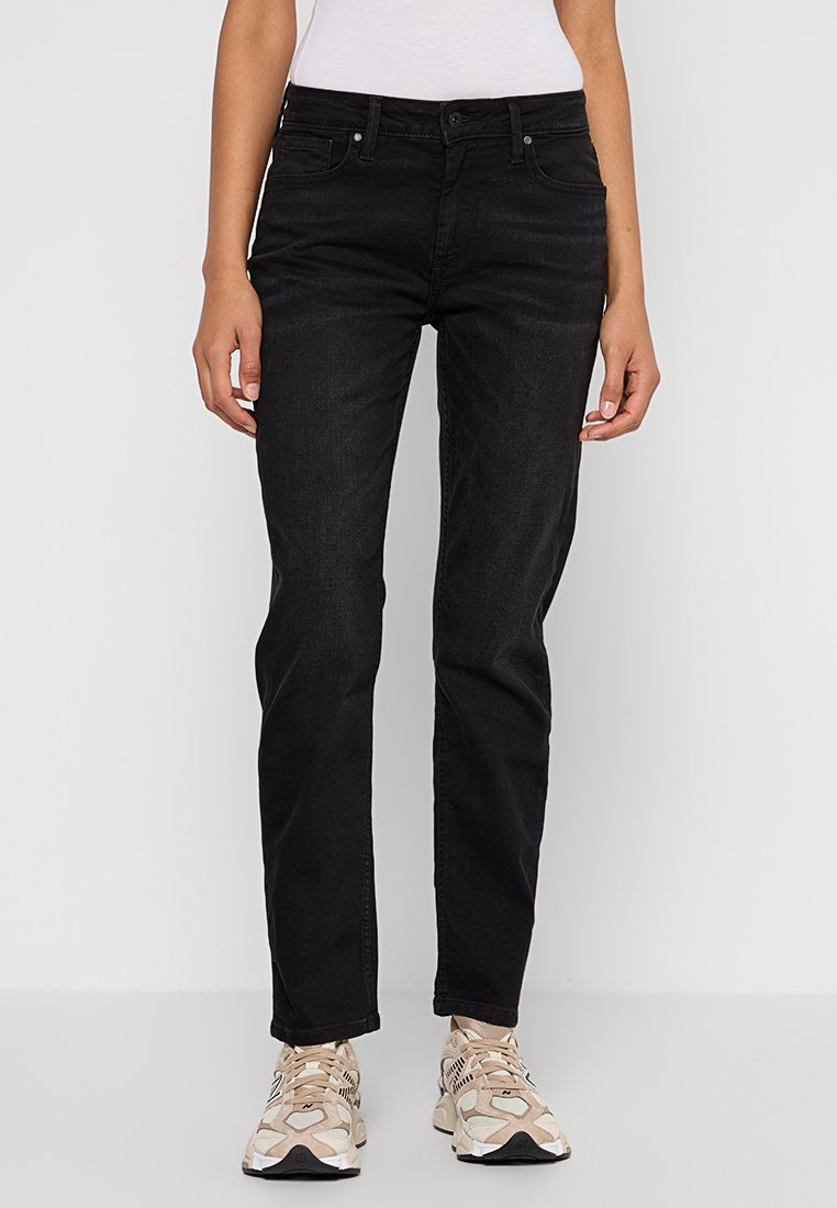 Femme portant un jean droit noir, un haut blanc et des baskets beiges à motifs, se tenant devant un fond blanc uni.