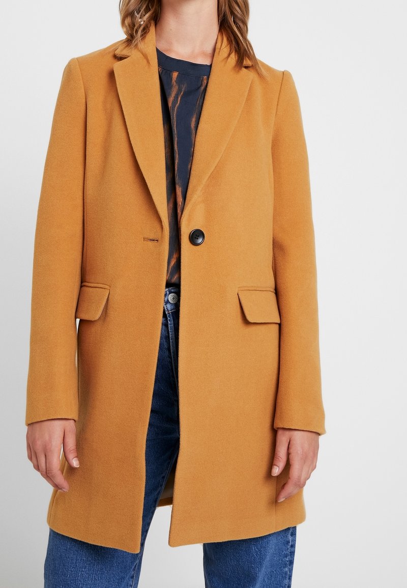 Femme portant un manteau camel à un seul bouton sur une chemise à motifs noir et marron et un jean bleu, les mains détendues le long du corps.