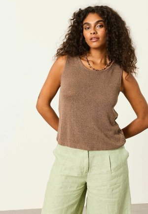 Woman with curly hair wearing a brown sleeveless knit top and light green linen pants, standing with hands behind back against plain background.