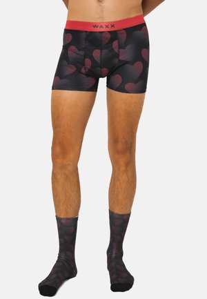 Homme portant un boxer noir et des chaussettes assorties avec des motifs de cœurs rouges et une ceinture rouge portant l'inscription "WAXX."