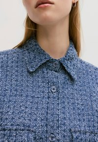 Blauwe textuurshirt met een kraag, voorzien van donkere knopen en een subtiel patroon. De stof lijkt zacht en geweven.